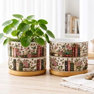 Può includere: Due vasi cilindrici in ceramica con finitura craquelé, caratterizzati da un motivo colorato di libri impilati, rose rosa e farfalle. Ogni vaso poggia su una base circolare in legno chiaro. Un vaso contiene una pianta Pilea verde.