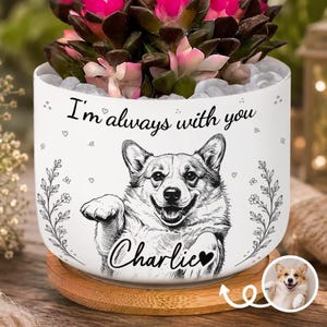 Pode incluir: Um vaso de cerâmica branca com uma ilustração de linhas pretas de um cão Corgi, o texto "I'm always with you" e o nome "Charlie" com um coração. O vaso assenta numa base circular de madeira e contém uma planta suculenta.