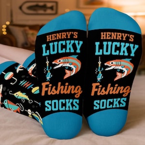 May include: A pair of black socks with blue cuffs and heels. The text reads HENRY'S LUCKY Fishing SOCKS in orange and blue. A graphic of a fish on a hook is centered between the text. The lower part of the socks features a pattern of fishing lures.
