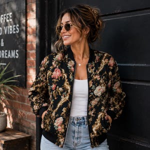 May include: A black bomber jacket featuring a detailed floral and ornate gold scrollwork pattern, a ribbed black collar, cuffs, and hem, and a front zipper closure, worn over a plain white top and light blue denim jeans.