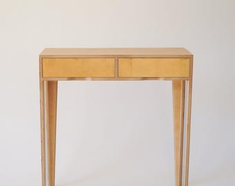 May include: A light wood console table with two drawers. The table has a simple, modern design with straight lines and a light wood finish.