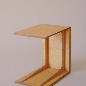 Puede incluir: Una mesa auxiliar de madera clara con un diseño único. La mesa tiene una tapa rectangular y una base curva.