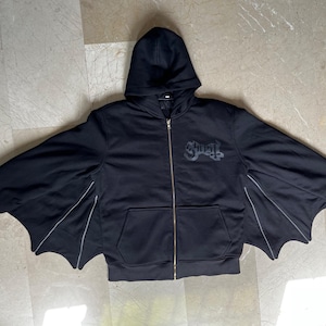 以下が含まれることがあります： 黒色のジップアップパーカー、コウモリの羽の袖付き。フード、フロントポケット、ゴールドカラーのジッパーが付いています。「Ghost」の文字が胸にダークグレーでプリントされています。袖はコウモリの羽のようにデザインされています。