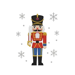 Puede incluir: Diseño bordado de un soldado cascanueces con uniforme rojo, detalles dorados, sombrero negro y espada. El diseño está rodeado de copos de nieve sobre un fondo blanco, ideal para manualidades navideñas.