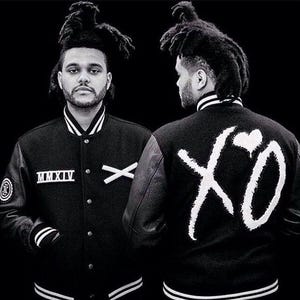 May include: Monochrome image of a person in a black varsity jacket with white trim. The jacket displays "MMXIV" and an "X" on the front, and a large "XO" graphic with a heart on the back. The person has dreadlocks and is facing the camera.