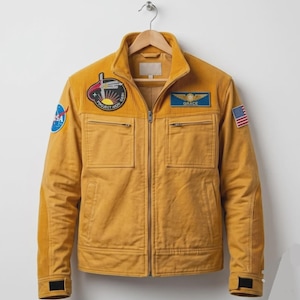 May include: A mustard yellow zip-up jacket featuring a ribbed collar and sleeve panels. The chest displays a Project Aiori patch and a Grace name tag. A NASA logo patch is on the right sleeve, and a United States flag patch is on the left sleeve.