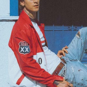 May include: A red and white color-blocked varsity jacket featuring a shield-shaped patch on the right sleeve with the text "lost XX" and a partial "0" visible on the front. The jacket is worn over a white t-shirt with blue jeans and a brown leather belt.