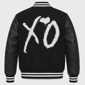 May include: A black varsity jacket featuring long black leather sleeves and a black wool-blend body. The back displays a large, white, textured chenille patch with the letters XO and a heart symbol. It has a ribbed collar and cuffs with white stripes.