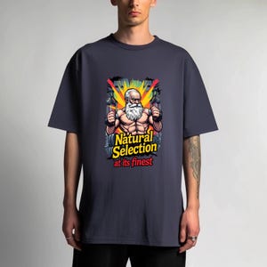 May include: Dark navy blue t-shirt featuring a cartoon illustration of a muscular, white-bearded man holding two objects. The graphic includes the text "Natural Selection at its finest" in yellow and red.