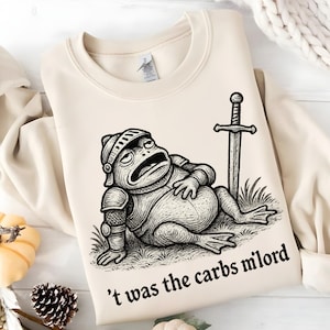 May include: Cream-colored sweatshirt featuring a cartoon frog in knight's armor, lying on grass next to a sword. The text "'t was the carbs milord" is printed below the frog. The sweatshirt has a crew neck and long sleeves.