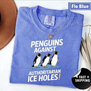 Peut inclure: Un t-shirt bleu avec l'inscription "PENGUINS AGAINST AUTHORITARIAN ICE HOLES!" et un dessin de trois pingouins. Le t-shirt a un col rond et des manches courtes. Un collier doré est posé sur le t-shirt.