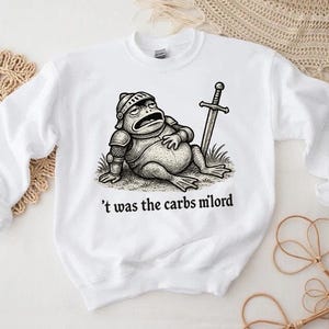 May include: White sweatshirt featuring a cartoon frog in knight's armor, resting with a sword. The phrase "'t was the carbs milord" is printed below the frog. The design is in black and white.