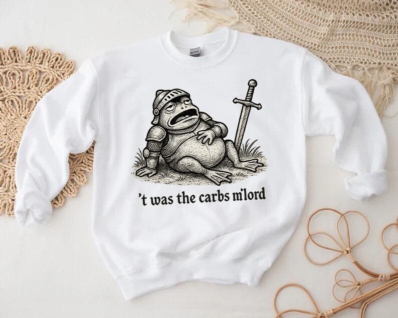 May include: White sweatshirt featuring a cartoon frog in knight's armor, resting with a sword. The phrase "'t was the carbs milord" is printed below the frog. The design is in black and white.