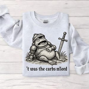 May include: Light gray sweatshirt featuring a cartoon frog in knight's armor, resting with a sword. The text "'t was the carbs milord" is printed below the frog. The sweatshirt has long sleeves and a crew neck.