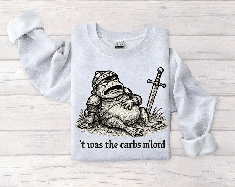 May include: Light gray sweatshirt featuring a cartoon frog in knight's armor, resting with a sword. The text "'t was the carbs milord" is printed below the frog. The sweatshirt has long sleeves and a crew neck.