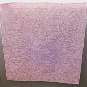 Puede incluir: Un edred&oacute;n rosa y blanco con un patr&oacute;n acolchado. El edred&oacute;n est&aacute; colgado en una pared. El edred&oacute;n est&aacute; hecho de un material suave y tiene una superficie texturizada. El edred&oacute;n tiene forma cuadrada.