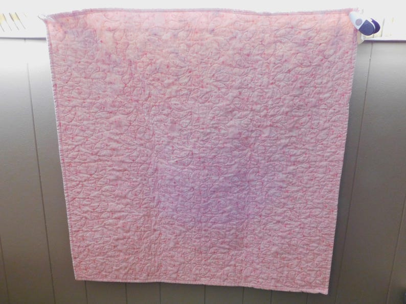 Puede incluir: Un edred&oacute;n rosa y blanco con un patr&oacute;n acolchado. El edred&oacute;n est&aacute; colgado en una pared. El edred&oacute;n est&aacute; hecho de un material suave y tiene una superficie texturizada. El edred&oacute;n tiene forma cuadrada.