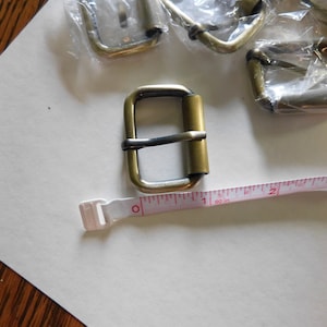 May include: Several brass-colored roller buckles, some still in plastic packaging, are displayed on a white surface. A white and pink measuring tape shows inches. One buckle is in focus, with a dark bar across the center.