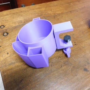 Könnte beinhalten: Ein violetter Getränkehalter aus Kunststoff mit einem runden Hauptfach, zwei kleineren quadratischen Ablagefächern und einem integrierten C-Klemmen-Mechanismus mit schwarzem Schraubknopf zur Befestigung an einer Tischkante.