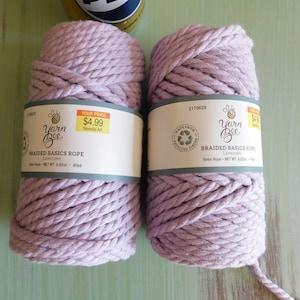 May include: Two spools of lavender-colored braided basics rope. Each spool has a label that reads "Yarn Bee Braided Basics Rope Lavender". The rope is 5mm in diameter and 40 yards in length. The label also includes a recycling symbol.