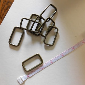 May include: A pile of eight gold-toned metal rectangular rings or loops, likely for attaching to a strap or belt. A white measuring tape with red markings is also in the image.