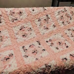 Puede incluir: Colcha rosa y blanca con un patr&oacute;n repetido de Minnie Mouse. La colcha tiene un dise&ntilde;o de patchwork con bloques cuadrados y un borde rosa. Las im&aacute;genes de Minnie Mouse son en negro, blanco y rosa.