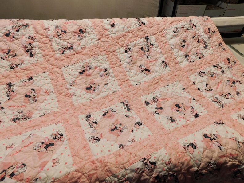 Puede incluir: Colcha rosa y blanca con un patr&oacute;n repetido de Minnie Mouse. La colcha tiene un dise&ntilde;o de patchwork con bloques cuadrados y un borde rosa. Las im&aacute;genes de Minnie Mouse son en negro, blanco y rosa.