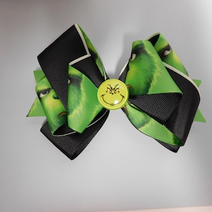 May include: A hair bow constructed from black and green patterned ribbon featuring a character face. The bow is layered with solid black ribbon and green ribbon printed with a green character, centred with a round yellow button displaying a character face.