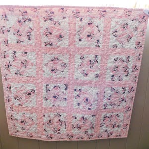 Puede incluir: Colcha rosa y blanca con un patr&oacute;n repetitivo de un personaje de dibujos animados. La colcha est&aacute; hecha de cuadrados, cada uno con el personaje sobre un fondo blanco, rodeado de rosa. La colcha est&aacute; colgada en una pared.