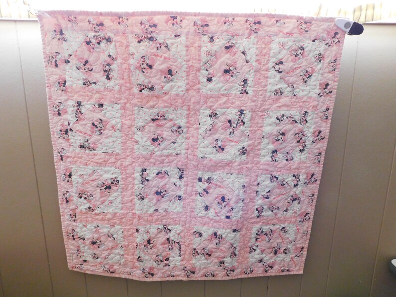 Puede incluir: Colcha rosa y blanca con un patr&oacute;n repetitivo de un personaje de dibujos animados. La colcha est&aacute; hecha de cuadrados, cada uno con el personaje sobre un fondo blanco, rodeado de rosa. La colcha est&aacute; colgada en una pared.