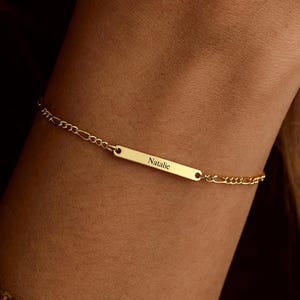 May include: A gold bracelet featuring a rectangular nameplate inscribed with "Natalie." The bracelet, with a delicate chain, is worn on a wrist. The jewellery item is gold in colour and is a piece of jewellery.