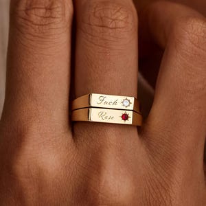 May include: Two gold rings stacked on a finger. The top ring has the word "Fuck" and a starburst design. The bottom ring has the word "Rose" and a small red gemstone. The rings are on a hand.