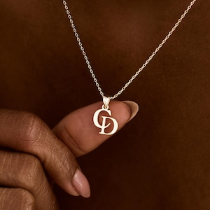 Peut inclure: Un collier doré avec une chaîne délicate et un pendentif représentant les lettres entrelacées "CD". Le pendentif est tenu par les doigts d'une personne. Le collier est un bijou.