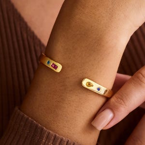 May include: Gold-coloured cuff bracelet with colourful gemstones. The open cuff design features a rectangular shape with a gemstone at each end. The gemstones are blue, red, and yellow. The bracelet is worn on a wrist.