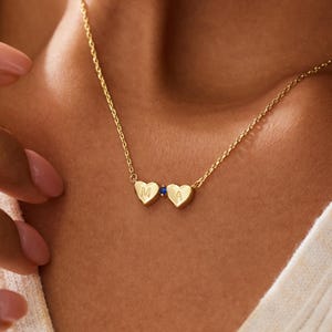 Op de afbeelding: Een goudkleurige ketting met twee kleine hartvormige bedels, elk gegraveerd met een enkele letter, M en A, gescheiden door een kleine blauwe edelsteen. De fijne ketting rust tegen de hals en toont de gepolijste afwerking.