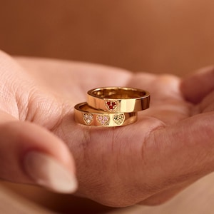 May include: Two stacked gold-toned rings featuring heart-shaped embellishments. The top ring displays a single red heart, while the bottom ring features three smaller heart-shaped accents in shades of pink, light pink, and yellow.