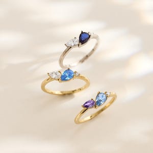 May include: Three rings featuring pear-cut and round gemstones in blue, white, and purple. The rings have gold and silver bands. The rings are displayed on a light-coloured surface.