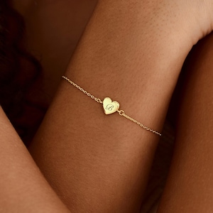 May include: A delicate gold chain bracelet featuring a small, centred heart-shaped charm. The heart charm is engraved with the uppercase letter B in a cursive script font. The bracelet is constructed with fine, polished gold links.