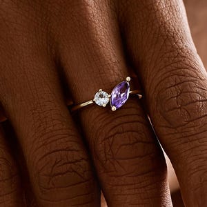 Peut inclure: Une bague en or ornée de deux pierres précieuses : une pierre ronde et transparente et une améthyste violette en forme de marquise. La bague est portée sur une main aux ongles manucurés, mettant en valeur le design et le contraste de couleurs.