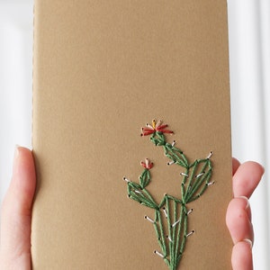 Puede incluir: Un cuaderno de color beige con un diseño de cactus bordado a mano. El cactus es verde con flores rojas y amarillas, y costuras blancas. El cuaderno está sostenido en las manos de una persona.