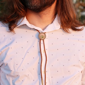 May include: A bolo tie with a circular, gold-toned pendant featuring a cactus design. The pendant is attached to a brown braided leather cord with gold-colored metal tips. The tie is worn over a white button-down shirt with a small, dark blue pattern.