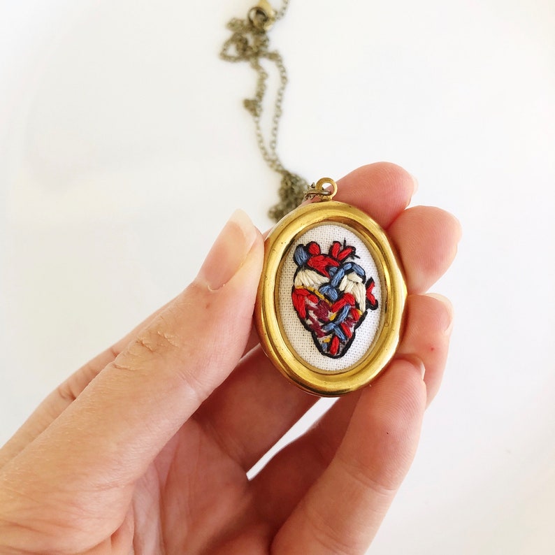 May include: A gold pendant necklace with a round frame featuring an embroidered anatomical heart in red, blue, and white.