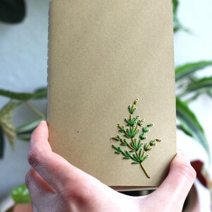 May include: A brown notebook with a green and yellow embroidered sprig of foliage on the front cover.
