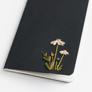 May include: A black notebook with a hand-stitched floral design. The embroidery features white daisies, green stems, and small colorful accents. The notebook is open, revealing blank pages.