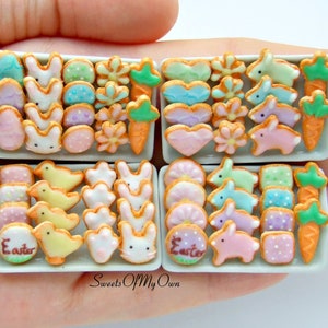 May include: Four white trays filled with miniature Easter-themed sugar cookies. The cookies are decorated with pastel colours and feature designs like bunnies, carrots, flowers, and eggs. The trays are labelled "Easter" and are arranged in a hand.