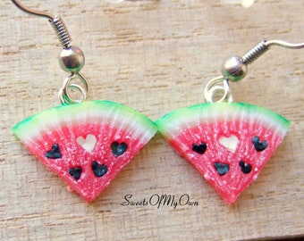 May include: A pair of watermelon slice earrings. Each earring features a red and white watermelon slice with a green rind, black heart-shaped seeds, and a white heart-shaped center. The earrings have silver-colored hooks.
