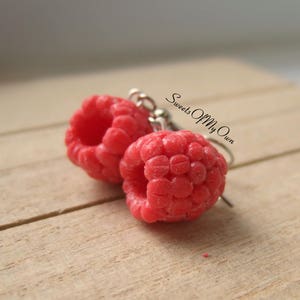 May include: A pair of red raspberry earrings with silver hooks. The earrings have a textured surface, resembling real raspberries. The earrings are set against a light brown wooden surface.