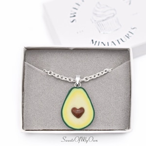 May include: A silver chain necklace with a pendant shaped like a halved avocado with a brown heart in the centre. The necklace is in a white box with grey lining. The box has the text "Sweets Of My Own Miniatures" on the top.