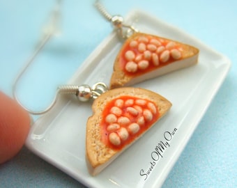 May include: A pair of earrings shaped like toast with baked beans on top. The earrings are on a white plate with the text "Sweets Of My Own" printed on the plate.