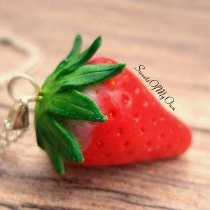 May include: A red and green polymer clay strawberry pendant on a silver chain. The strawberry has a white tip and a green leafy top. The pendant is labeled "Sweets Of My Own".